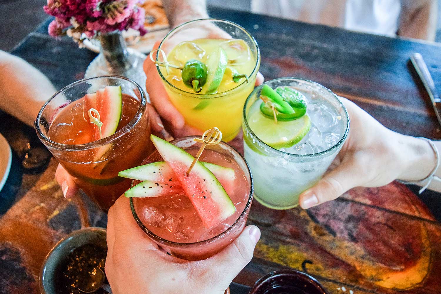 Four hands holding assorted cocktail drinks, cheers-ing each other.