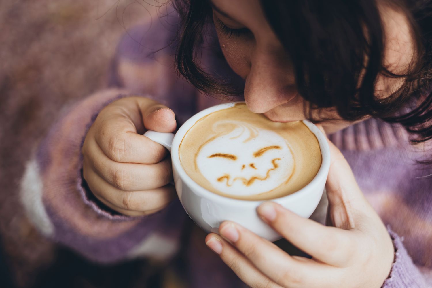 Person drinking a cup of coffee with latte art, wearing a purple sweater.
