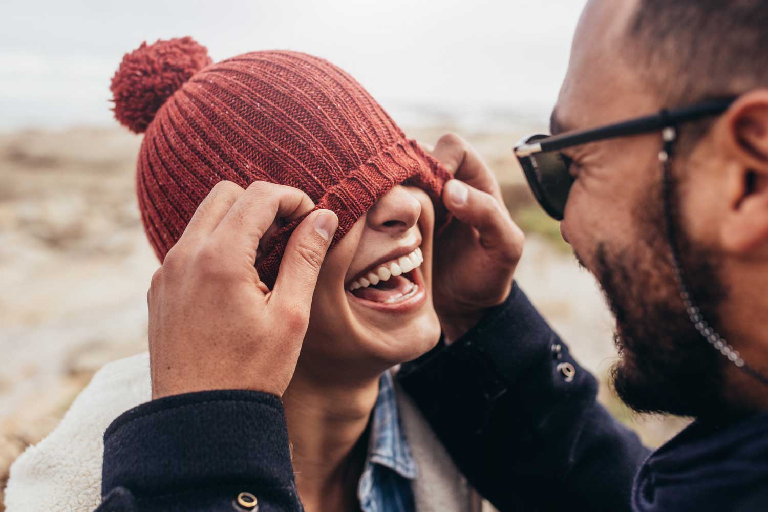 Attractive couple laughing, man red pulls beanie over girl's eyes.