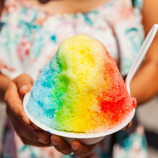 Close-up of woman's hands holding Hawaiian rainbow shave ice, indicating flavor of "Hawaii Five-OH" flavored lip balm by EXOH.